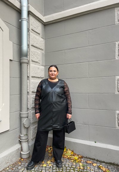 Vestido de piel sintética negra superpuesto sobre una blusa negra de encaje de mangas largas, combinado con pantalones negros de pierna ancha y un bolso pequeño. Fondo de pared de piedra.