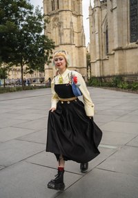 Persona con cabello corto teñido viste un vestido negro, una camisa amarilla, un cinturón de cadena dorada y botas gruesas, caminando sobre un pavimento de piedra urbana cerca de un edificio histórico de piedra.