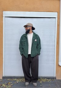 Homme portant une veste en velours côtelé verte, un pull beige, un pantalon noir et une casquette, debout les mains dans les poches devant un volet métallique blanc fermé.