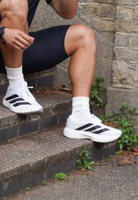Osoba w czarnych szortach i białych trampkach Adidas z czarnymi paskami, siedząca na zewnętrznych kamiennych schodach z kilkoma zielonymi roślinami w pobliżu.