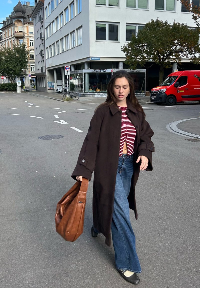 Casaco de lã castanho, top curto às riscas vermelhas e brancas, jeans flare de ganga, a carregar uma mala de couro castanha, a caminhar numa rua da cidade.