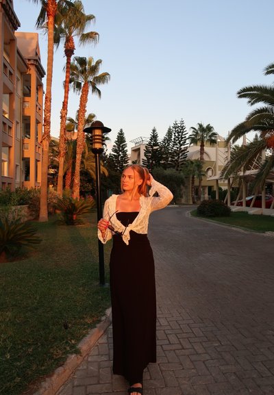 Vestido maxi negro con una cintura ajustada y cárdigan de encaje. De pie en un camino adoquinado, rodeada de palmeras y edificios modernos.