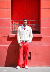 Πλεκτό κρεμ ζακέτα με πλέξεις καλωδίων, κόκκινη γραβάτα και φαρδιά κόκκινα παντελόνια. Το μοντέλο στέκεται μπροστά σε έναν κόκκινο τοίχο και διακοσμητική περίφραξη.