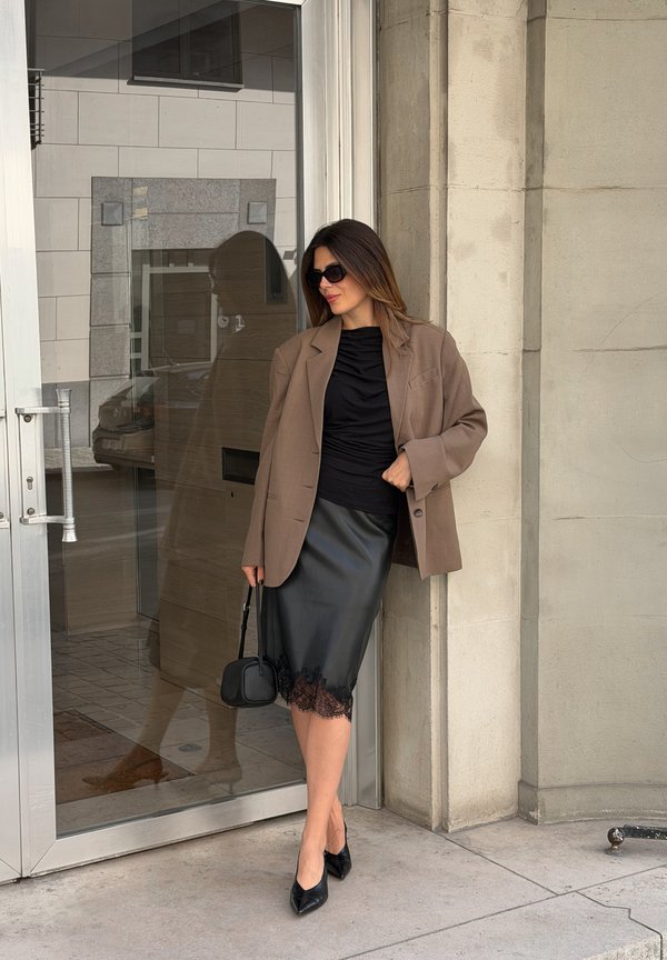 Mujer con gafas de sol, blazer marrón, top negro fruncido, falda negra con encaje y tacones apoyada contra una pared de piedra junto a una puerta de cristal.