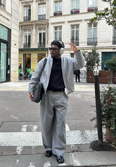 Chaqueta bomber gris sobre un suéter negro, pantalones grises anchos, zapatos negros y auriculares blancos. Gafas de sol oscuras y una bolsa negra. Entorno urbano.