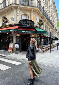 Musta nahkatakki, oliivinvihreä maksimekko korkealla halkiolla, mustat saappaat. Taustalla kahvila vihreällä markiisilla ja neonkyltillä.