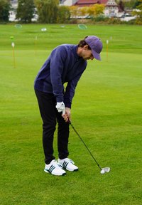 Golfimängija tumesinise triibulise pika varrukaga särgi ja mustade pükstega, kasutades klubipulka valge golfipalli kõrval rohelisel murul. Valged kingad roheliste detailidega.