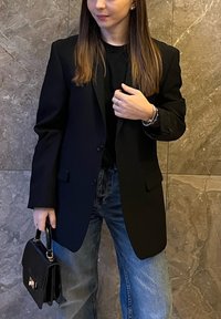 Czarna, dopasowana marynarka wykonana z gładkiego materiału, o klasycznym kroju z jednorzędowym zapięciem i klapami z wycięciem. Połączona z niebieskimi jeansami.