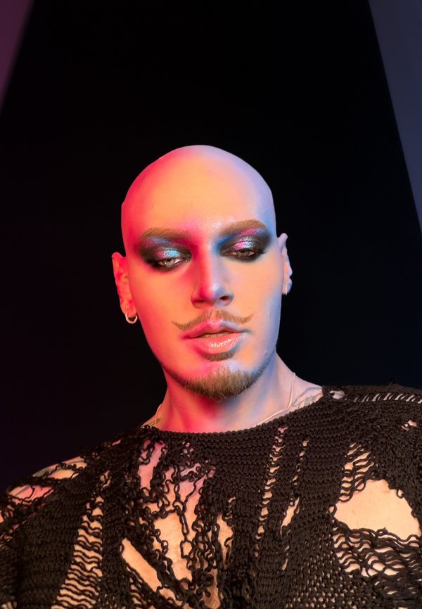 Skaldet person med stylet overskæg og skæg bærer glitrende, røgfarvet øjen make-up og en sort, åbenstrikket top mod en mørk baggrund med farverig belysning.