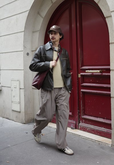 Chaqueta de cuero marrón sobre suéter beige, pantalones grises holgados y zapatillas beige con rayas negras. Lleva un bolso burdeos, caminando cerca de una puerta roja.