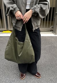 Ramenní taška z lesklé měkké kůže olivově zelené barvy se sklady a velkým otvorem. Nese ji osoba v černých kalhotách a hnědých botách.