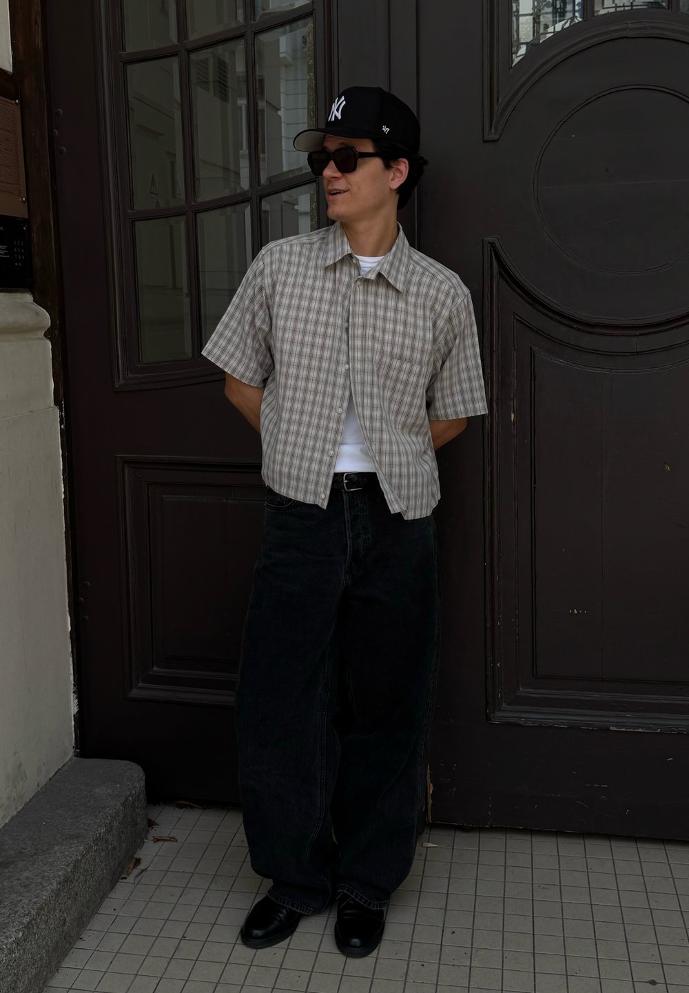Uomo in piedi con le mani dietro la schiena, indossa un cappellino nero, occhiali da sole, camicia a quadri a maniche corte, canottiera bianca, pantaloni neri larghi e scarpe nere.