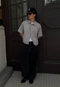 Uomo in piedi con le mani dietro la schiena, indossa un cappellino nero, occhiali da sole, camicia a quadri a maniche corte, canottiera bianca, pantaloni neri larghi e scarpe nere.