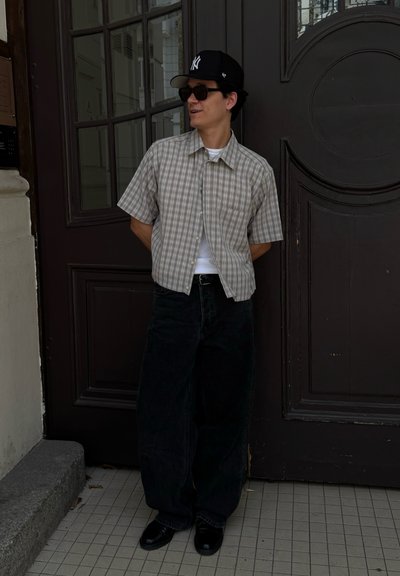 Hombre de pie con las manos detrás de la espalda, usando una gorra negra, gafas de sol, una camisa de cuadros de manga corta, una camiseta blanca por debajo, pantalones anchos negros y zapatos negros.