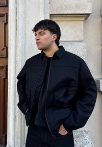 Jeune homme aux cheveux foncés portant une veste noire et un pull, debout les mains dans les poches contre un mur en pierre texturé et une porte en bois.