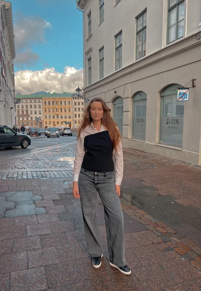 Camisa blanca de botones debajo de un chaleco negro ajustado, combinada con pantalones vaqueros grises de pierna ancha y tenis negros, de pie en una calle de adoquines.