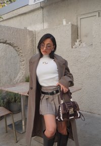 Manteau marron oversize sur un top blanc côtelé. Mini-jupe plissée à carreaux avec ceinture noire. Sac à main marron avec détails en mesh et ferrures dorées.