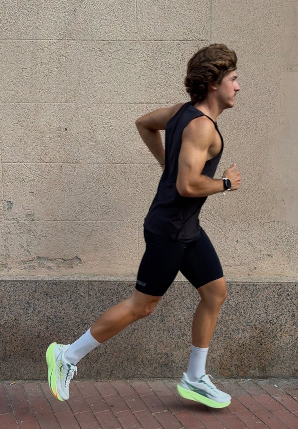 Giovane uomo in abbigliamento sportivo nero che corre su un marciapiede di mattoni accanto a un muro beige con texture.