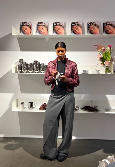 Chaqueta de cuero burdeos, top negro y pantalones de pierna ancha grises. Copas de plata y un arreglo floral expuestos en estanterías blancas.