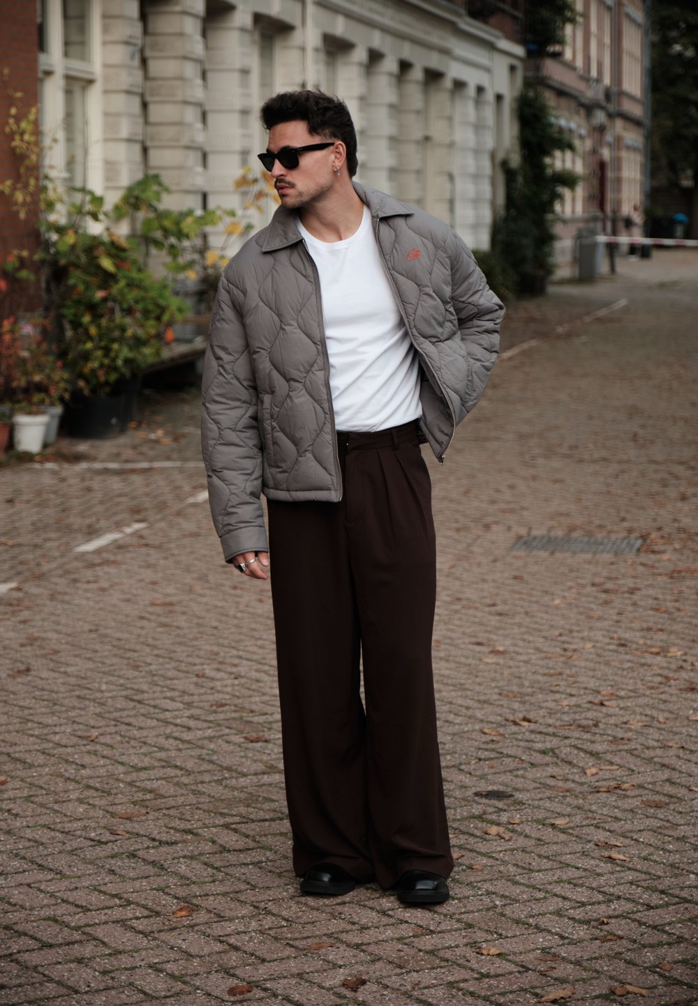 Veste grise matelassée avec un col, t-shirt blanc et large pantalon brun. Des lunettes de soleil noires et des chaussures noires élégantes complètent le look.