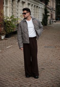 Veste grise matelassée avec un col, t-shirt blanc et large pantalon brun. Des lunettes de soleil noires et des chaussures noires élégantes complètent le look.