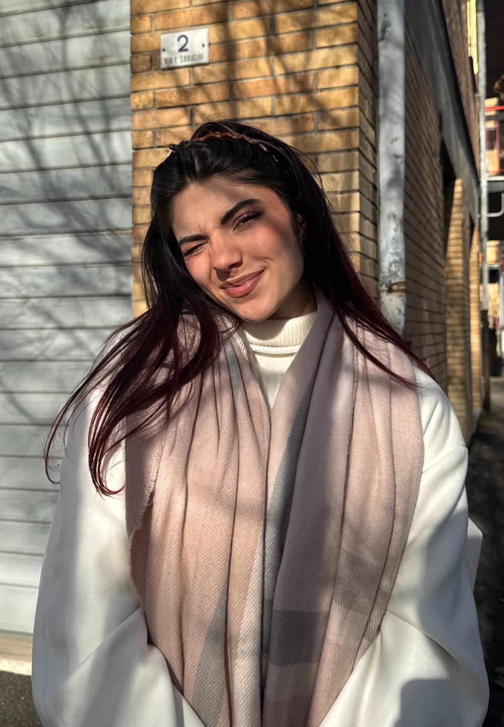 Ljusrosa och grå mönstrad halsduk draperad över en vit tröja, med långt mörkt hår och solljus som kastar skuggor på en tegelvägg.