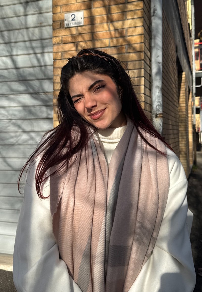 Hellrosa und grau gemusterter Schal, der über einen weißen Pullover gelegt ist, mit langen dunklen Haaren und Sonnenlicht, das Schatten auf eine Backsteinmauer wirft.