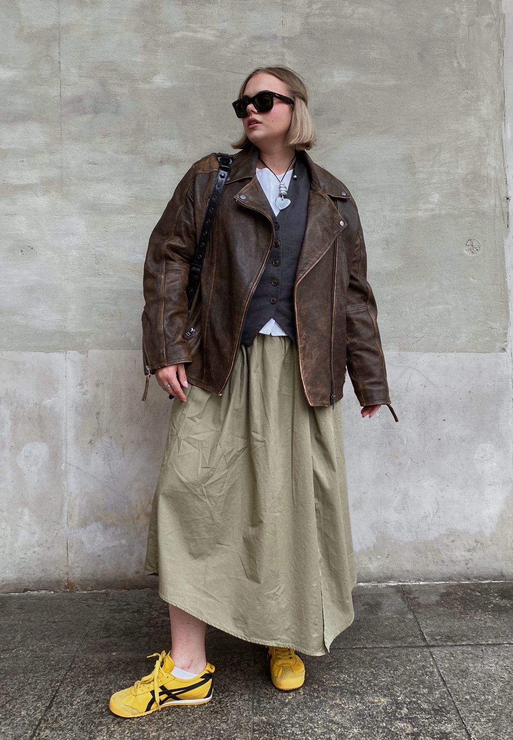 Καφέ δερμάτινο μπουφάν μοτοσικλετιστή, γκρι γιλέκο, λευκό πουκάμισο, ελιωτό πράσινο φούστα και κίτρινες αθλητικές σόλες με μαύρες λεπτομέρειες. Απαλές υφές και στρωτή δομή.