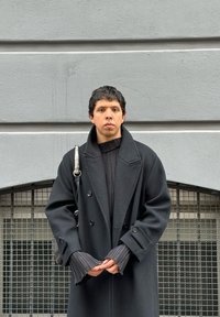 Μαύρο διπλό-κόφτη παλτό με φαρδιές πέτες, συνδυασμένο με ριμπ μαύρο πουλόβερ και υφασμάτινα μανίκια; περιλαμβάνει μεταλλική ζώνη ώμου.