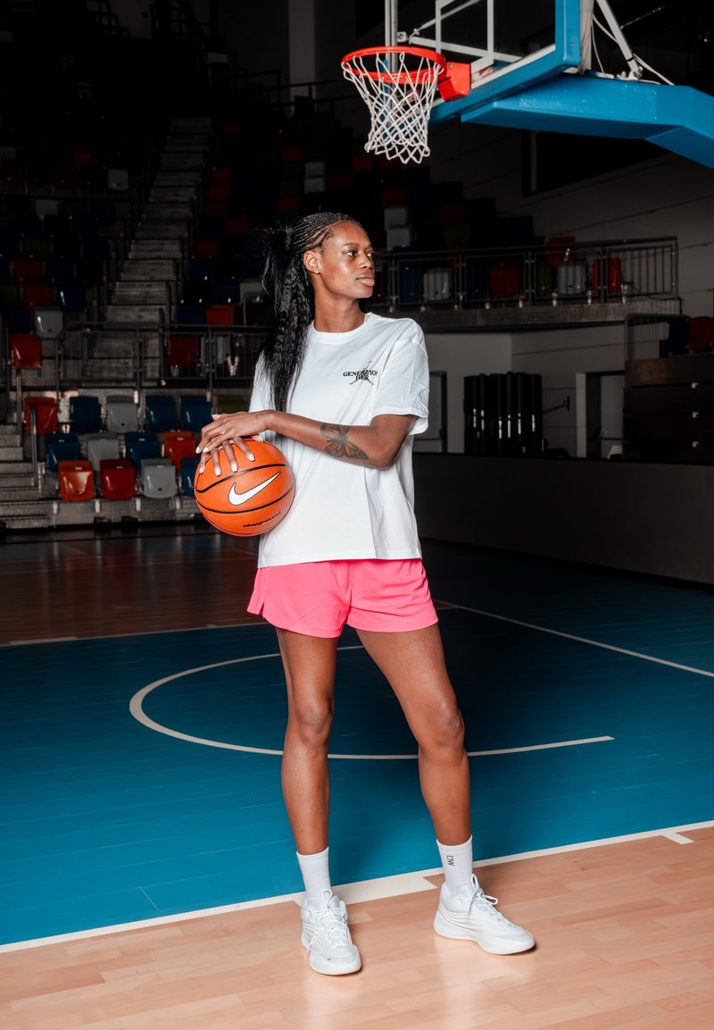 Mujer con camisa blanca y pantalones cortos rosas sosteniendo un baloncesto en una cancha cubierta bajo la canasta, mirando hacia la derecha.