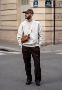 Grå genser, brun cap, brun crossbody-veske, svarte bukser og sko. Mine detaljer inkluderer lagdelte halskjeder og teksturert stoff.