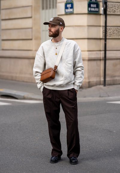 Sudadera gris, gorra marrón, bolso cruzado marrón, pantalones negros y zapatos. Detalles notables incluyen collares en capas y tejido texturizado.