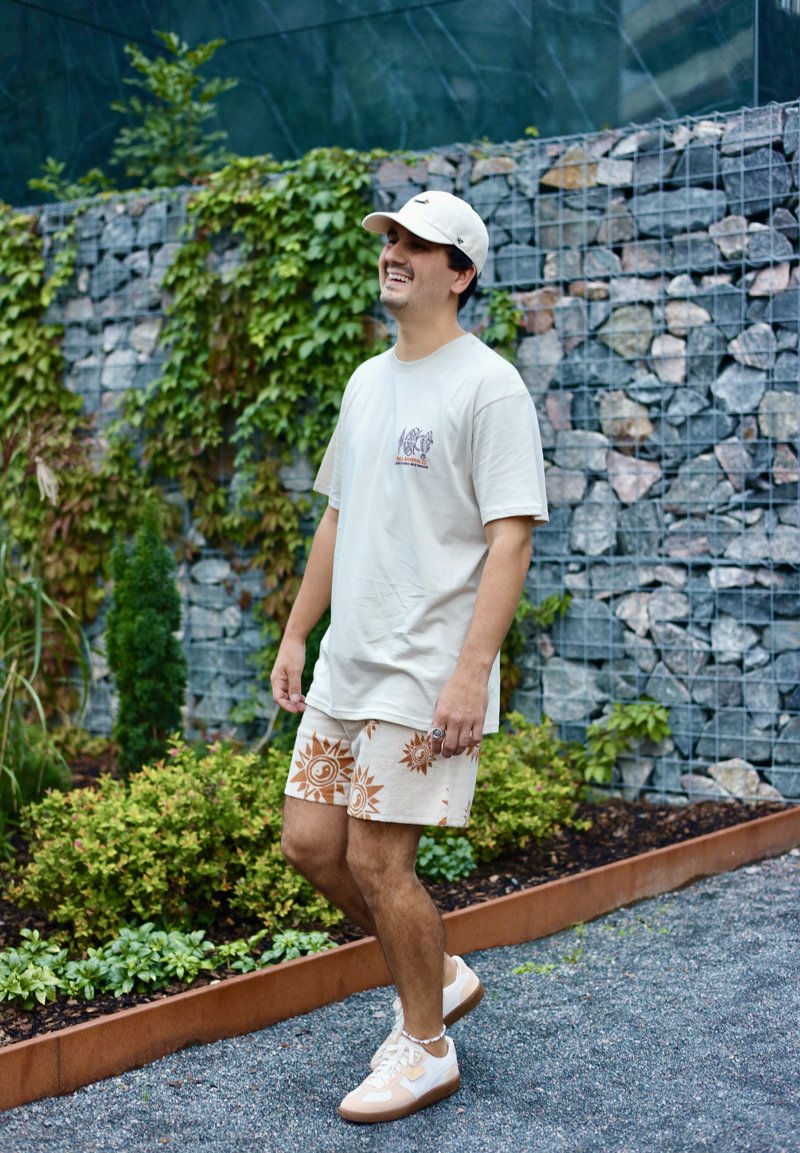 Beige T-shirt med grafik, ljusbruna shorts med solmotiv och vita sneakers med beige detaljer, mot en bakgrund av grönska och stenvägg.