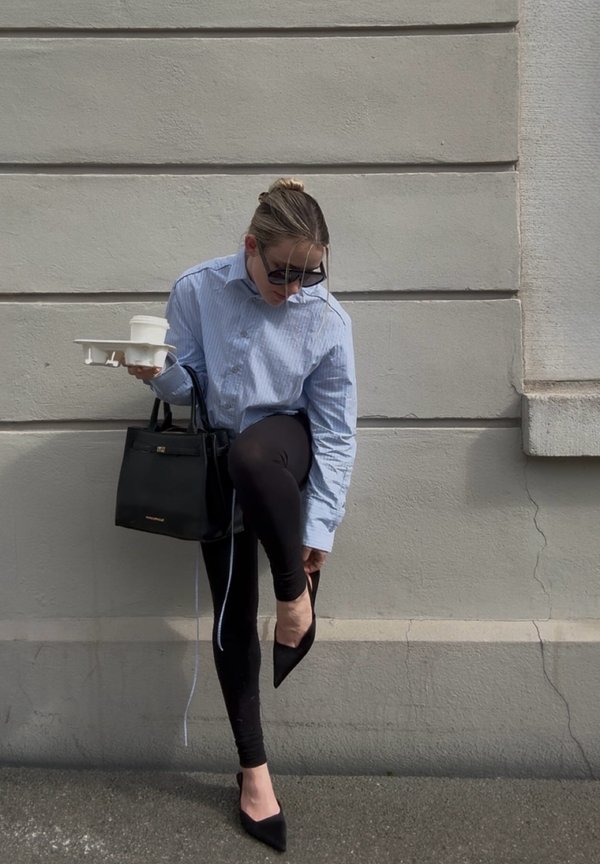 Kvinde med solbriller iført en blå skjorte og sorte leggings tager sorte spidse sko på, mens hun holder en sort håndtaske og en hvid drikkebærer.