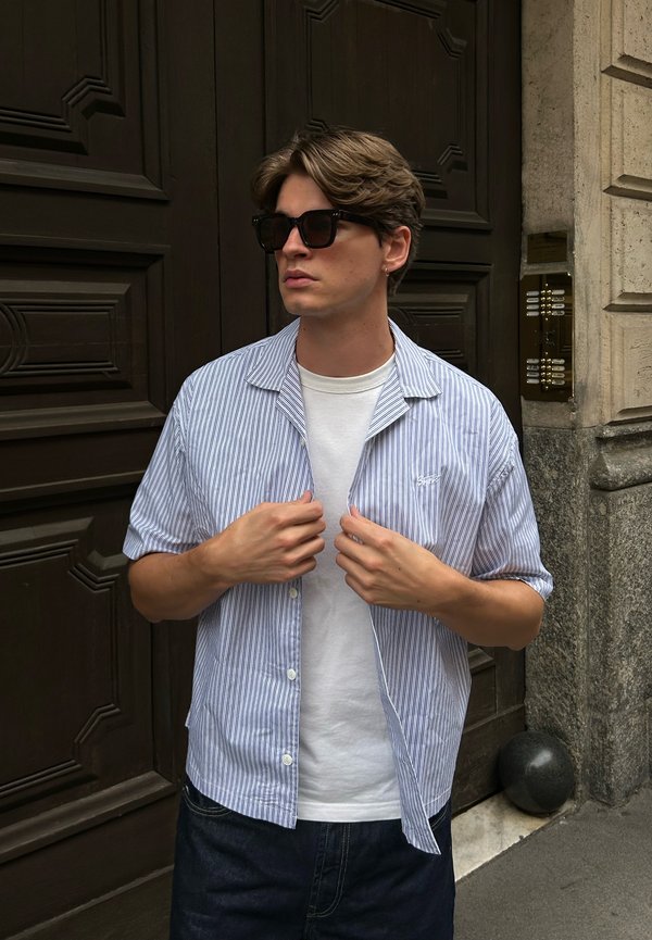 Chemise à manches courtes à boutons rayée bleu clair et blanche, portée sur un T-shirt blanc ; lunettes de soleil noires, shorts en denim décontractés.