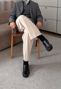 Homme assis en tailleur sur une chaise en bois, portant un blazer gris, un pull foncé, un pantalon clair, des chaussettes noires et des mocassins noirs dans un environnement intérieur.