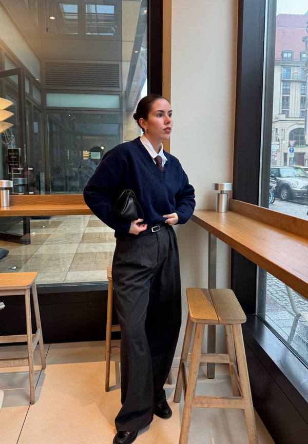 Ung kvinde i marineblå sweater og brede sorte bukser står inde i en café ved vinduet og holder en sort clutch, mens hun kigger udenfor.