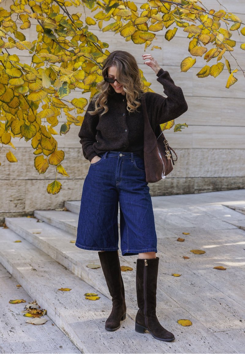 Maglione marrone, culotte di denim blu e stivali alti in camoscio marrone. Accessori includono occhiali da sole e una borsa a tracolla marrone. Foglie autunnali sullo sfondo.