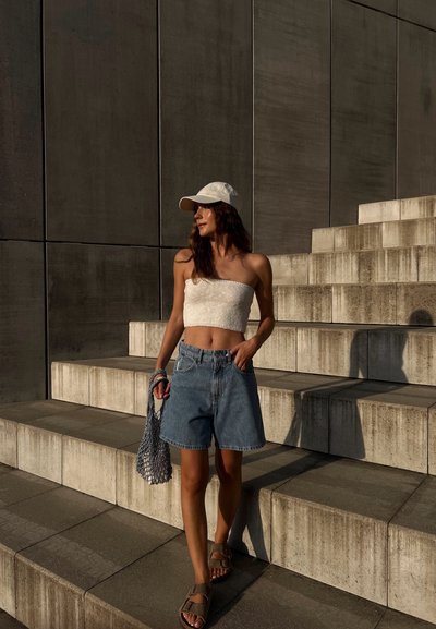 Top corto de algodón en blanco, pantalones cortos de mezclilla azul de cintura alta, sandalias color tostado y gorra de béisbol blanca; llevando una bolsa de malla, de pie sobre escaleras de piedra.