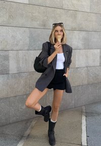 Blazer gris de doble botonadura, camiseta blanca sin mangas, pantalones cortos negros, botas altas negras con correas, sosteniendo un helado en un cono, de pie contra una pared de piedra.