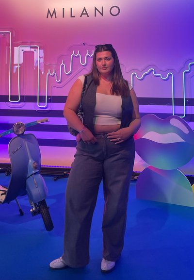 Scooter vintage azul junto a una persona que lleva un chaleco gris, una camiseta corta blanca y pantalones anchos grises. Fondo colorido con letreros de neón.