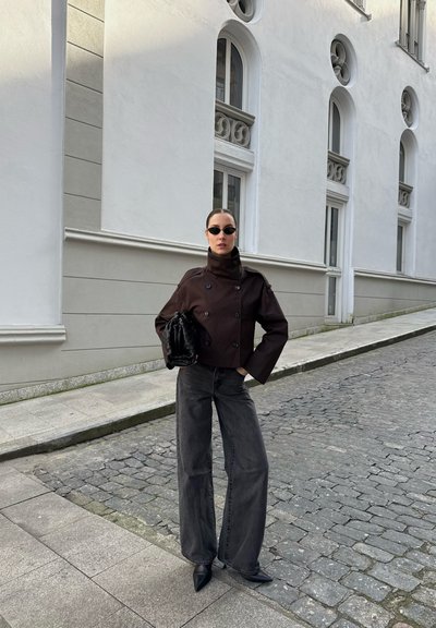 Mujer con chaqueta oscura de doble botonadura, vaqueros anchos negros, zapatos puntiagudos y gafas de sol pequeñas, de pie en una calle empedrada junto a un edificio blanco.