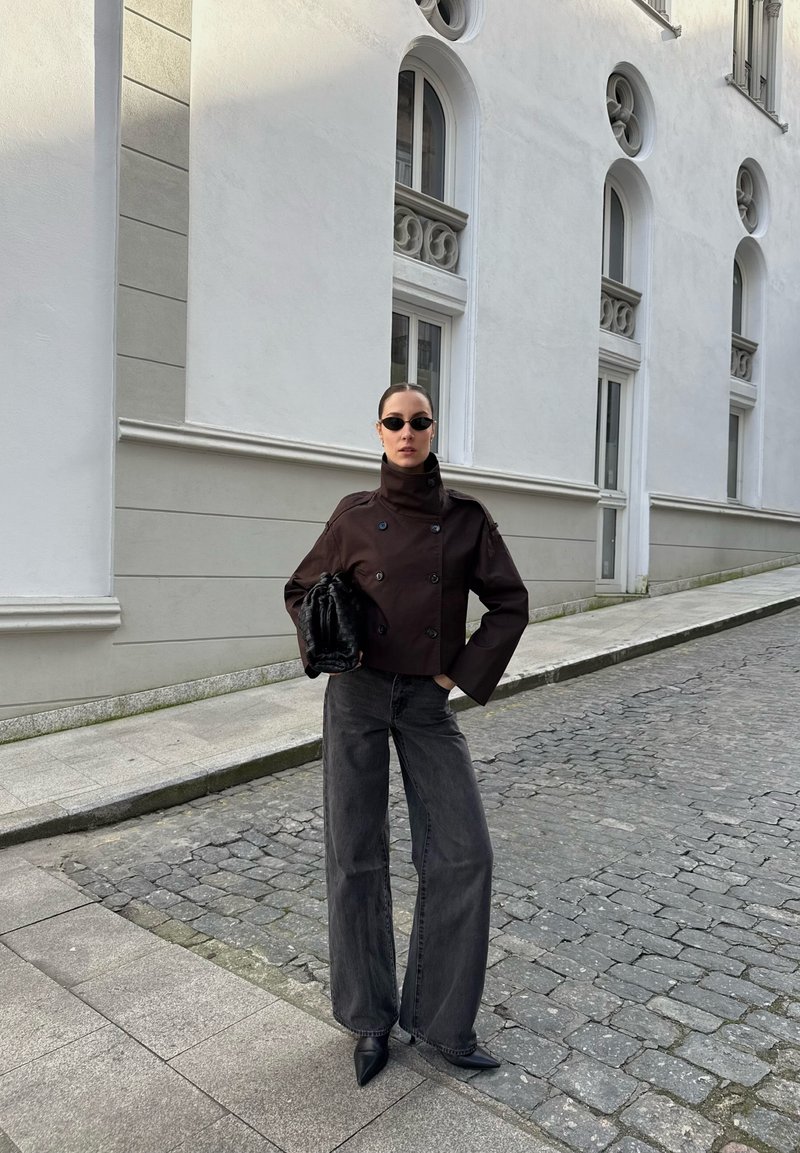Femme portant une veste sombre à double boutonnage, un large jean noir, des chaussures pointues et de petites lunettes de soleil, se tenant sur une rue pavée près d'un bâtiment blanc.