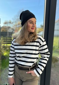 Ριγέ μπλούζα με μακριά μανίκια σε λευκό και μαύρο χρώμα, συνδυασμένη με μπεζ παντελόνι και μαύρο σκουφάκι. Το ύφασμα φαίνεται μαλακό και έχει άνετη εφαρμογή.