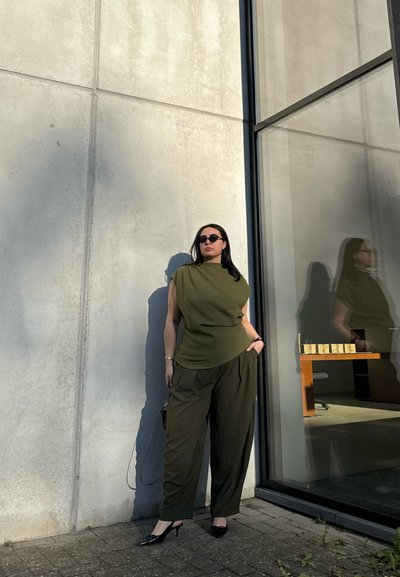 Mujer con top sin mangas y pantalones verde oliva, con gafas de sol, de pie junto a una pared de hormigón cerca de una ventana de vidrio con su reflejo visible.