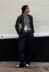 Woman with dark hair in a bun wearing a long black coat, a grey zip-up sweater, black trousers, a green belt, and a green handbag standing against a wall.
