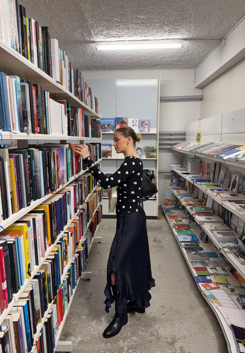 Femme portant un haut à pois noirs et une longue jupe, choisissant un livre sur une étagère haute dans une allée bien éclairée d'une librairie.