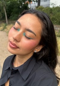 Retrato de una persona con los ojos cerrados, vistiendo una camisa negra. Presenta delineador de ojos en color aguamarina, mejillas sonrosadas y aretes plateados. Su cabello es oscuro y suelto.