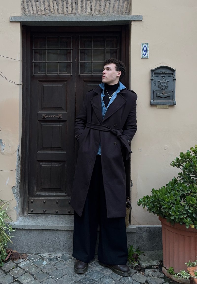 Homme portant un long manteau sombre et une veste en jean, debout les mains dans les poches près d'une porte en bois et d'une boîte aux lettres numérotée 9, avec des plantes en pot à proximité.