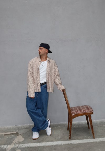 Conjunto casual que incluye una camisa de cuadros beige, una camiseta blanca, unos pantalones vaqueros azules holgados y zapatillas blancas; de pie junto a una silla de madera con un asiento estampado.